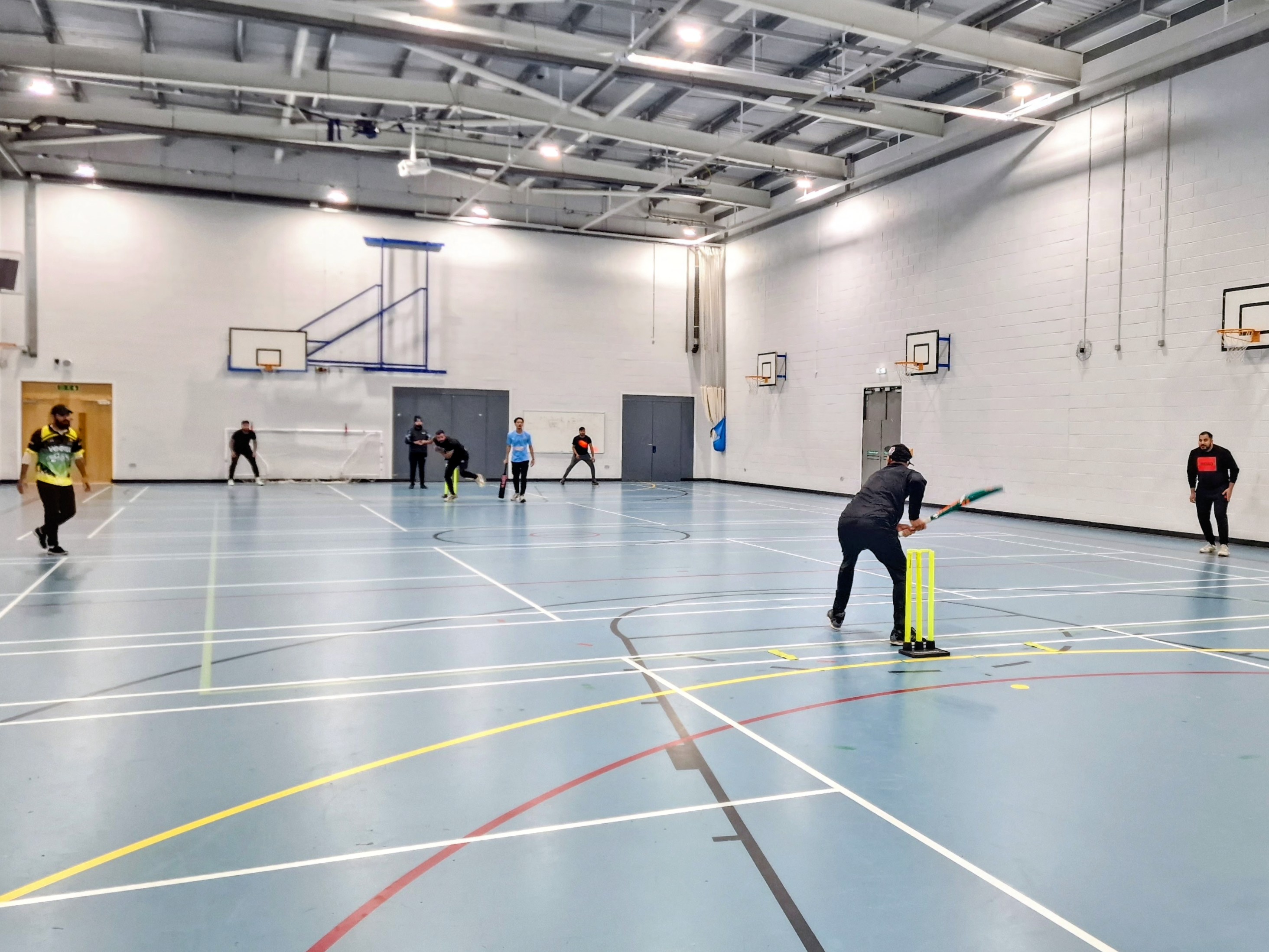 Wide view of a BBICL 2024 indoor cricket game at Sheffield Park Academy, showing both teams in position under the bright sports hall lights.