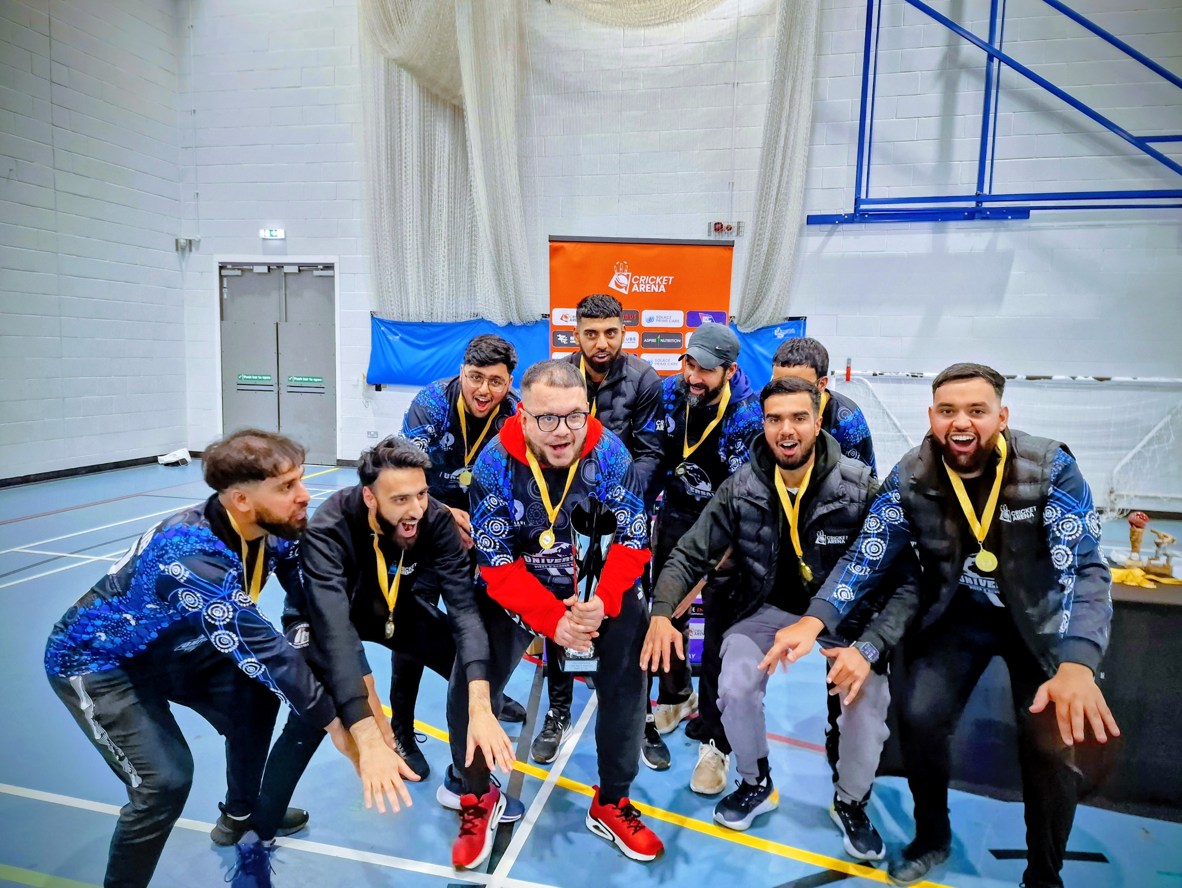 Darnall Dynamites crouch together with medals and trophy at BBICL 2024, shouting and smiling just before lifting the indoor league title in Sheffield.