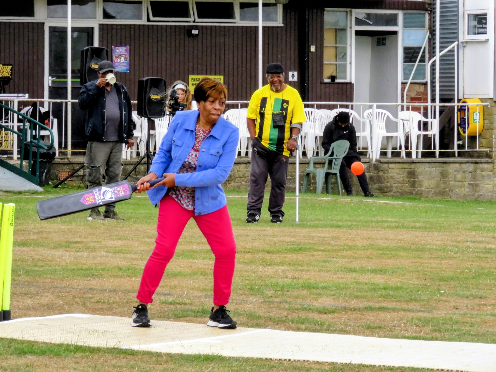 Woman batting in a friendly soft-ball cricket game on the outfield during Windrush Day celebrations.