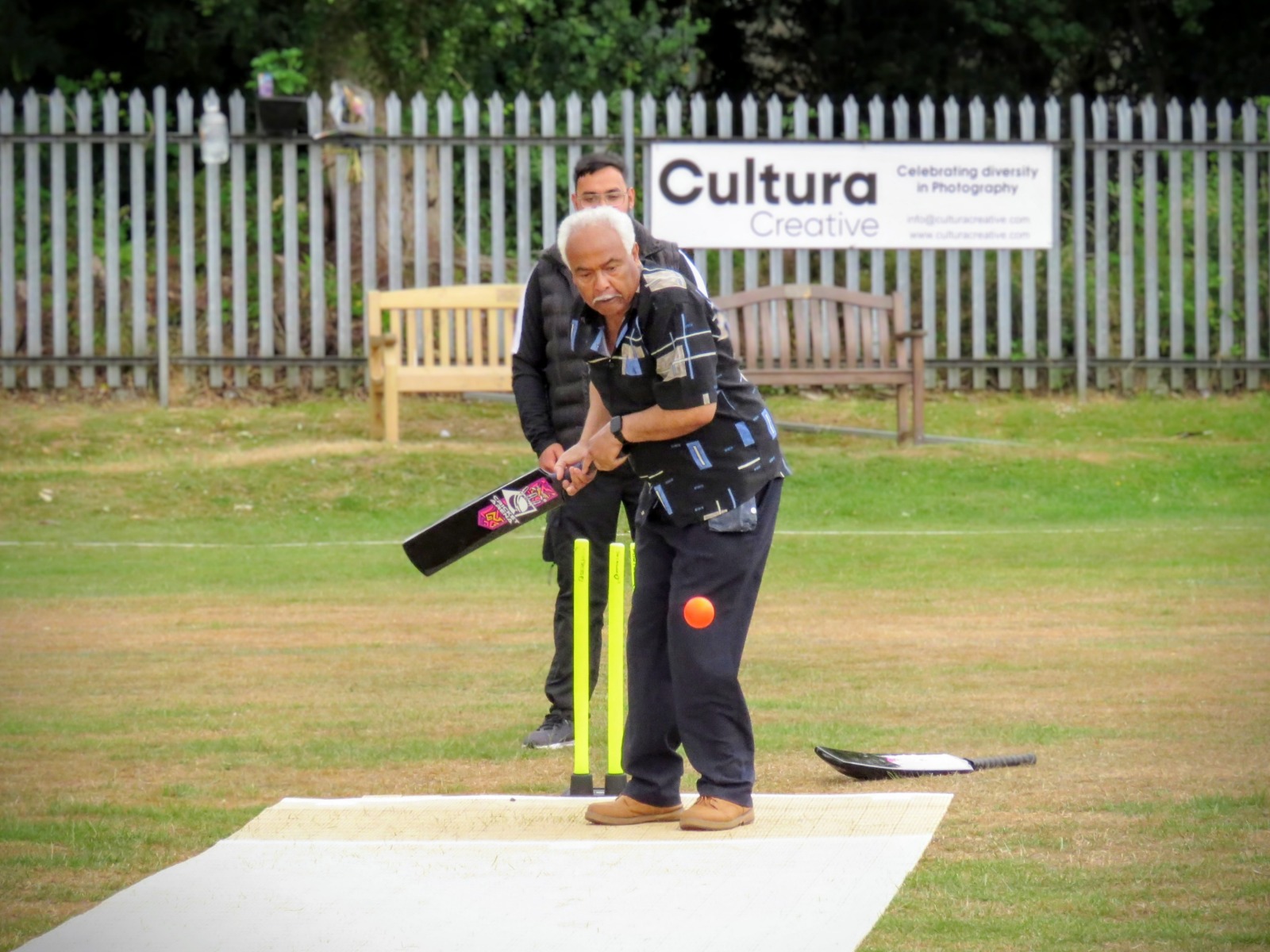 Older community member batting in a soft-ball cricket game at Caribbean Sports Club on Windrush Day 2025.