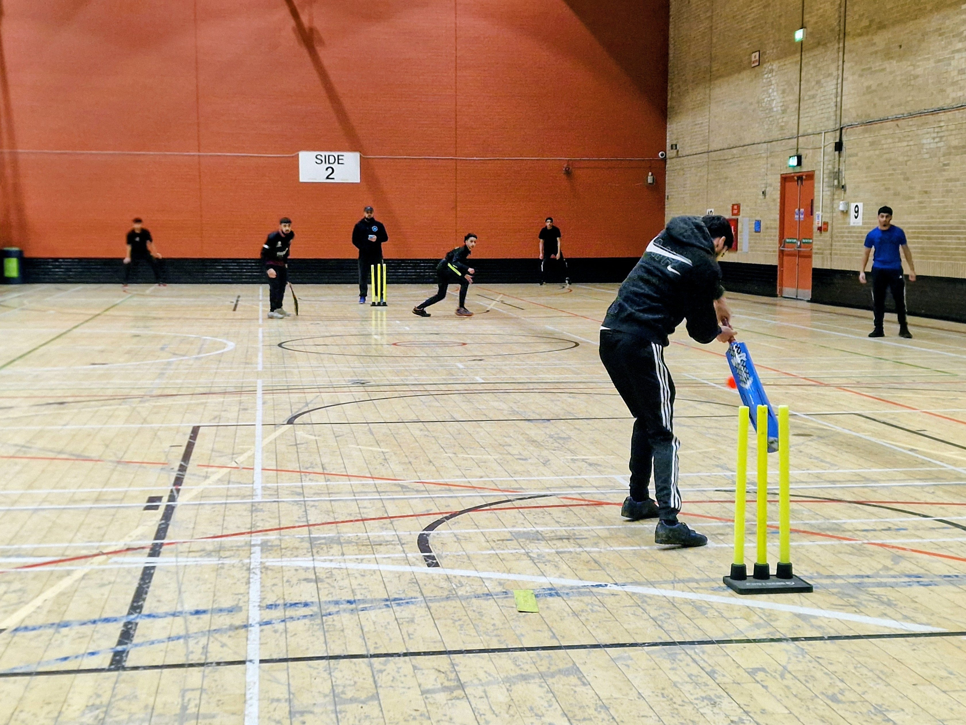Young players compete in an indoor windball cricket match at Concord Sports Centre in Sheffield, with a batter striking the ball toward bright yellow stumps.