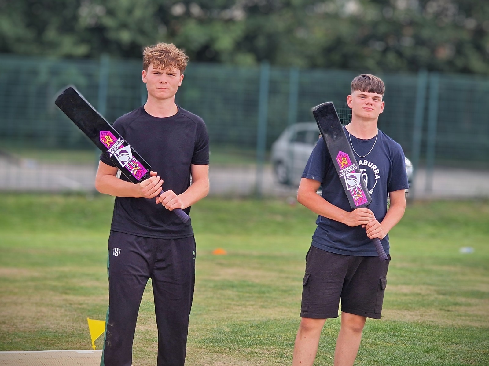 Two Junior Hundred 2025 participants stand on the outfield at Shiregreen Cricket Club holding Cricket Arena branded fibre bats used for the soft-ball tournament.