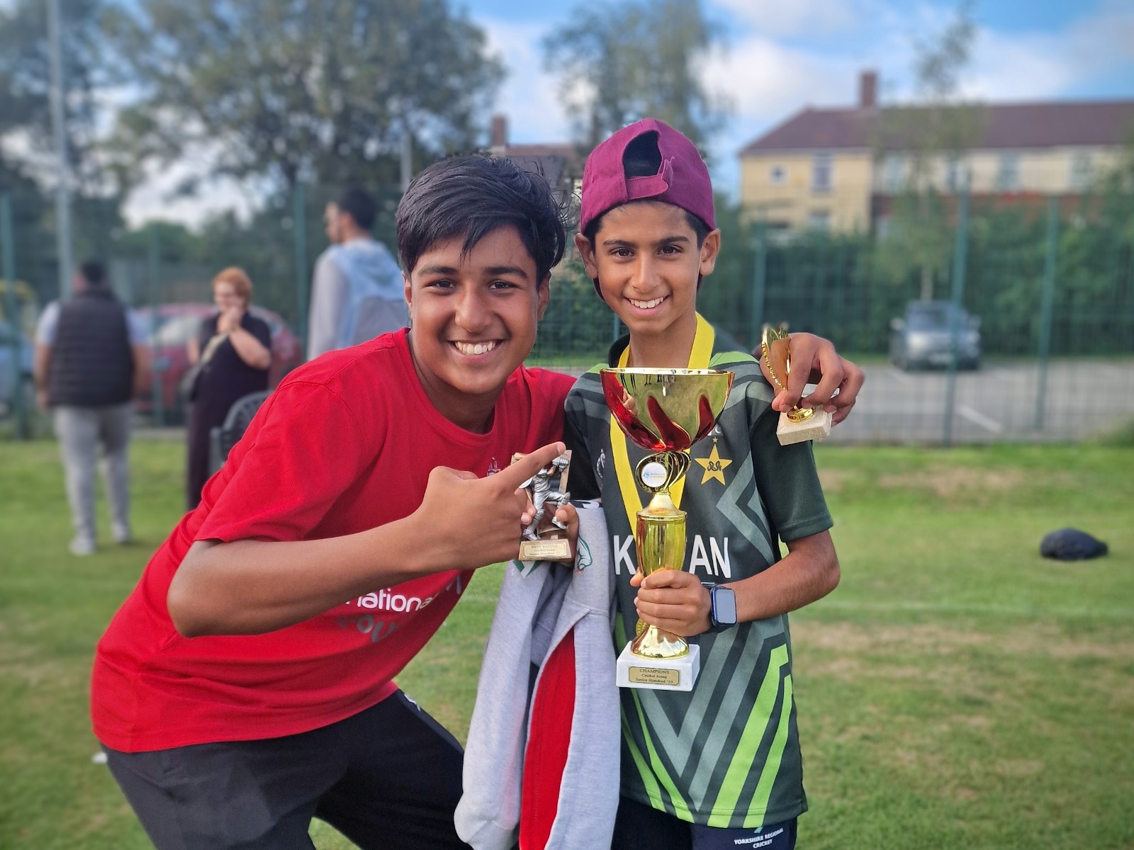The youngest Junior Hundred 2025 player celebrates with a friend, holding a winners’ trophy and medal on the outfield at Shiregreen Cricket Club in Sheffield.