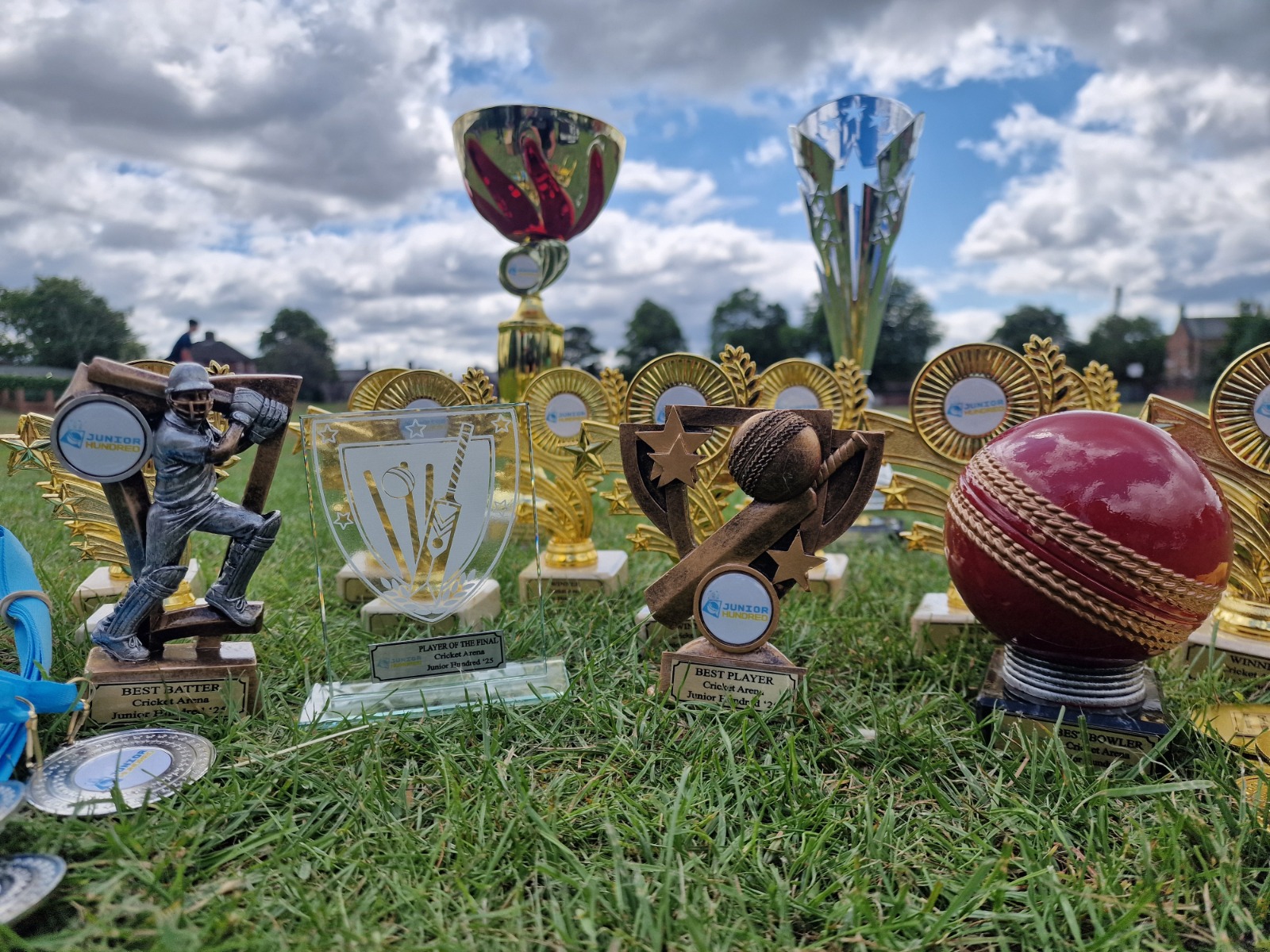 Close-up of Junior Hundred 2025 trophies and medals laid out on the grass at Shiregreen Cricket Club in Sheffield before the presentation.