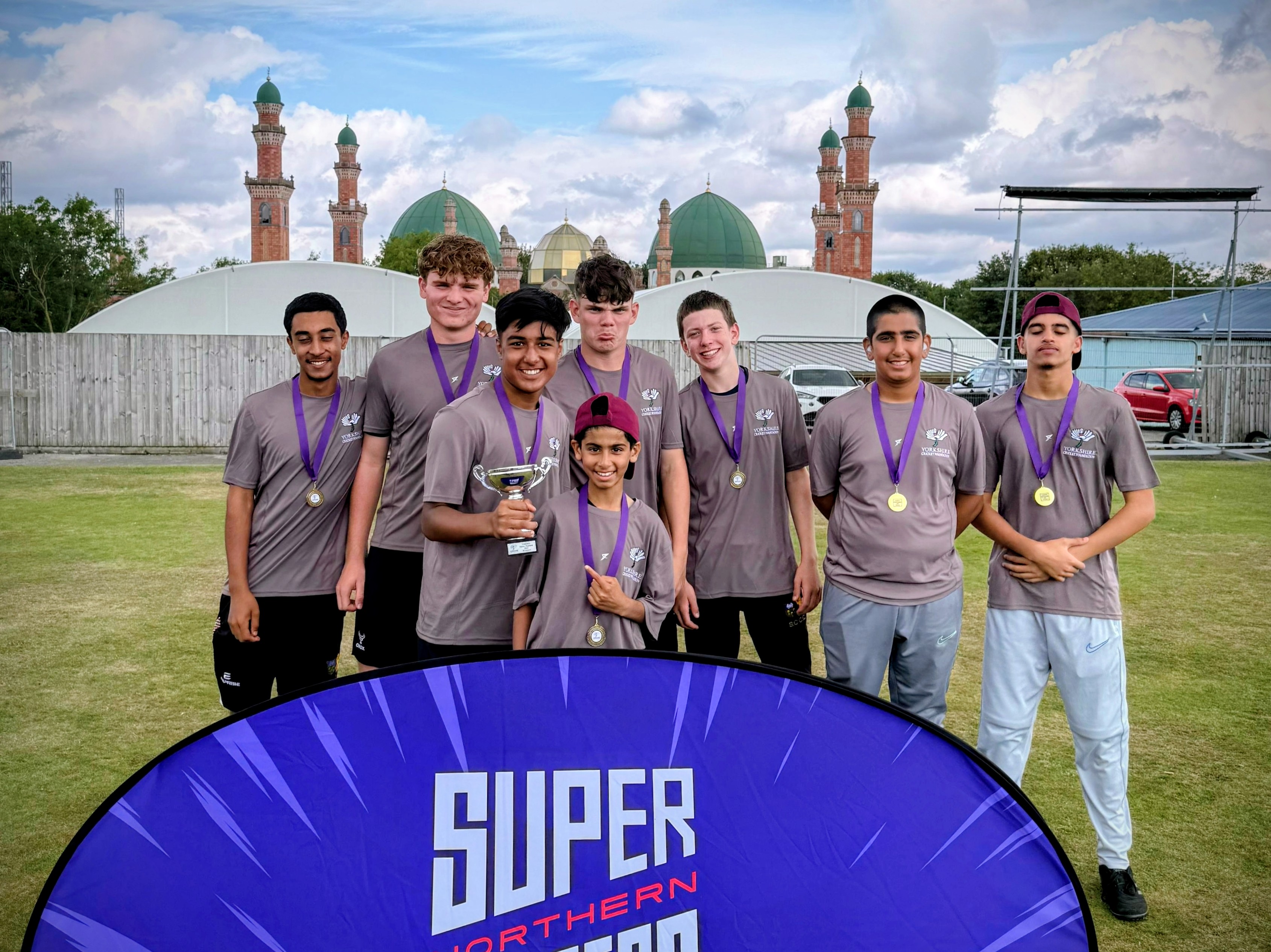 Sharrow Stars players pose with medals and the Yorkshire Junior Hundred trophy behind a Super Northern Chargers banner.