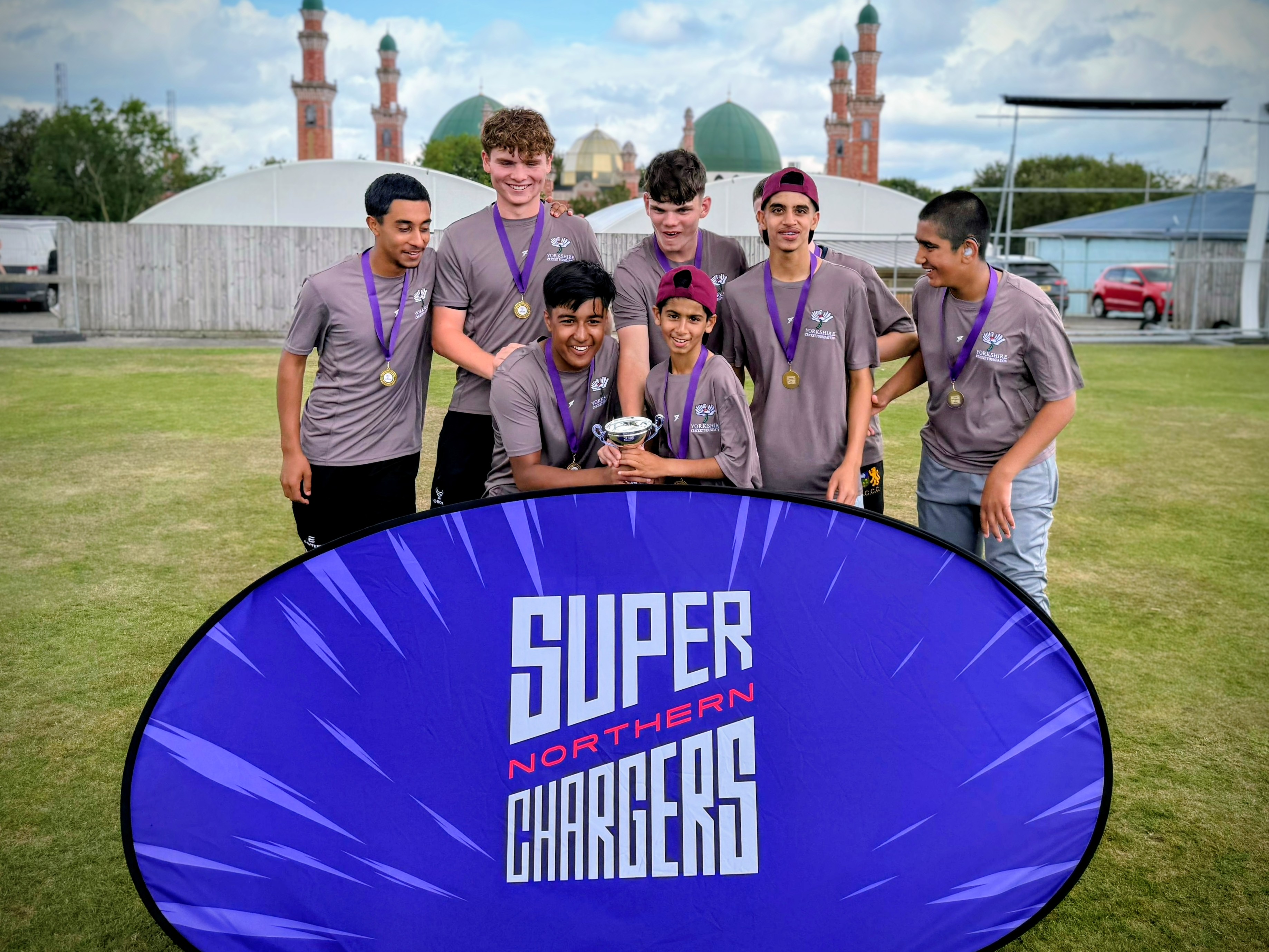 Sharrow Stars squad crouch behind a Super Northern Chargers banner with the Junior Hundred trophy at Park Avenue, Bradford.