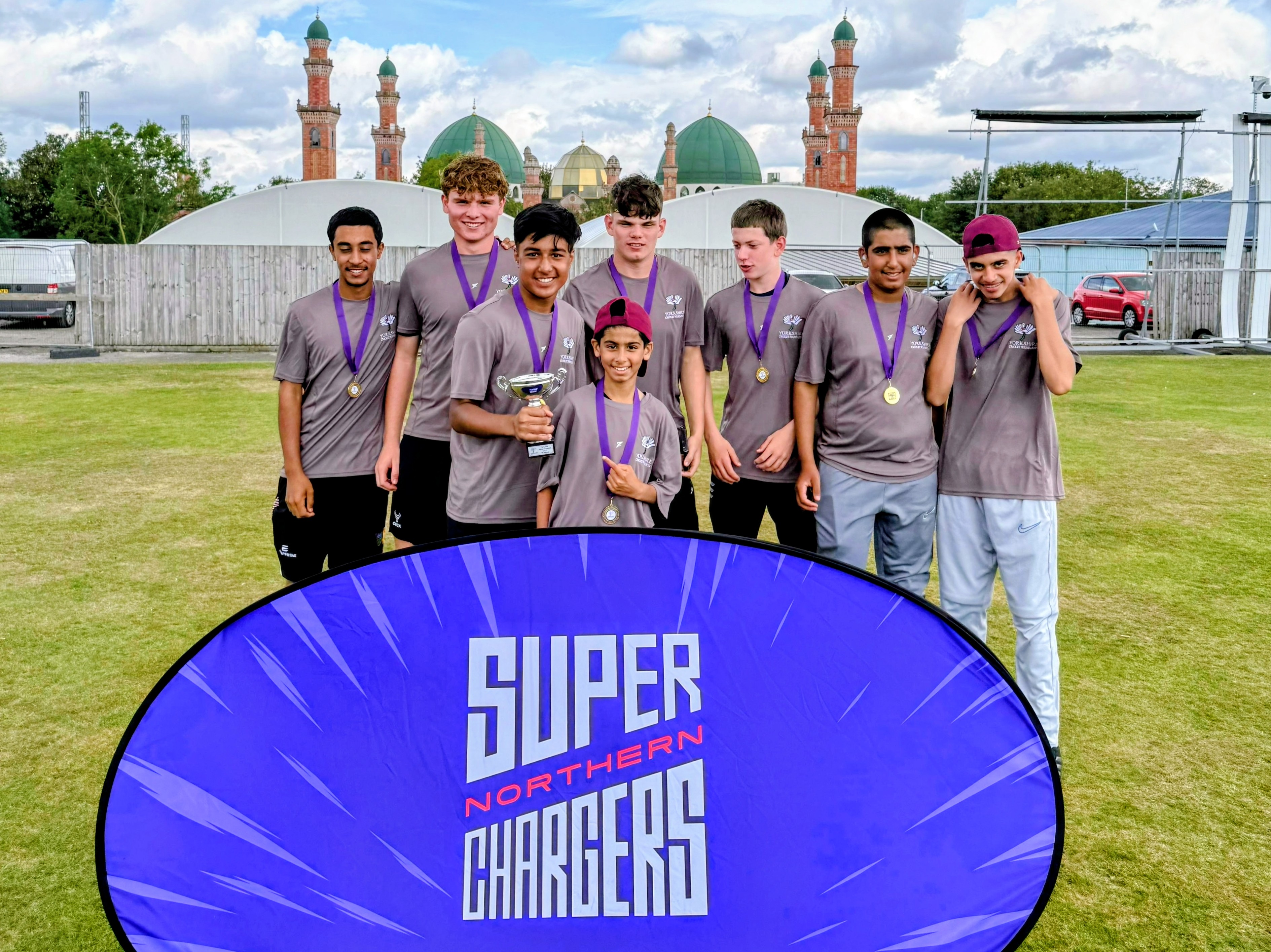 Sharrow Stars champions line up with the Junior Hundred trophy and medals behind a Super Northern Chargers banner at Park Avenue, Bradford.