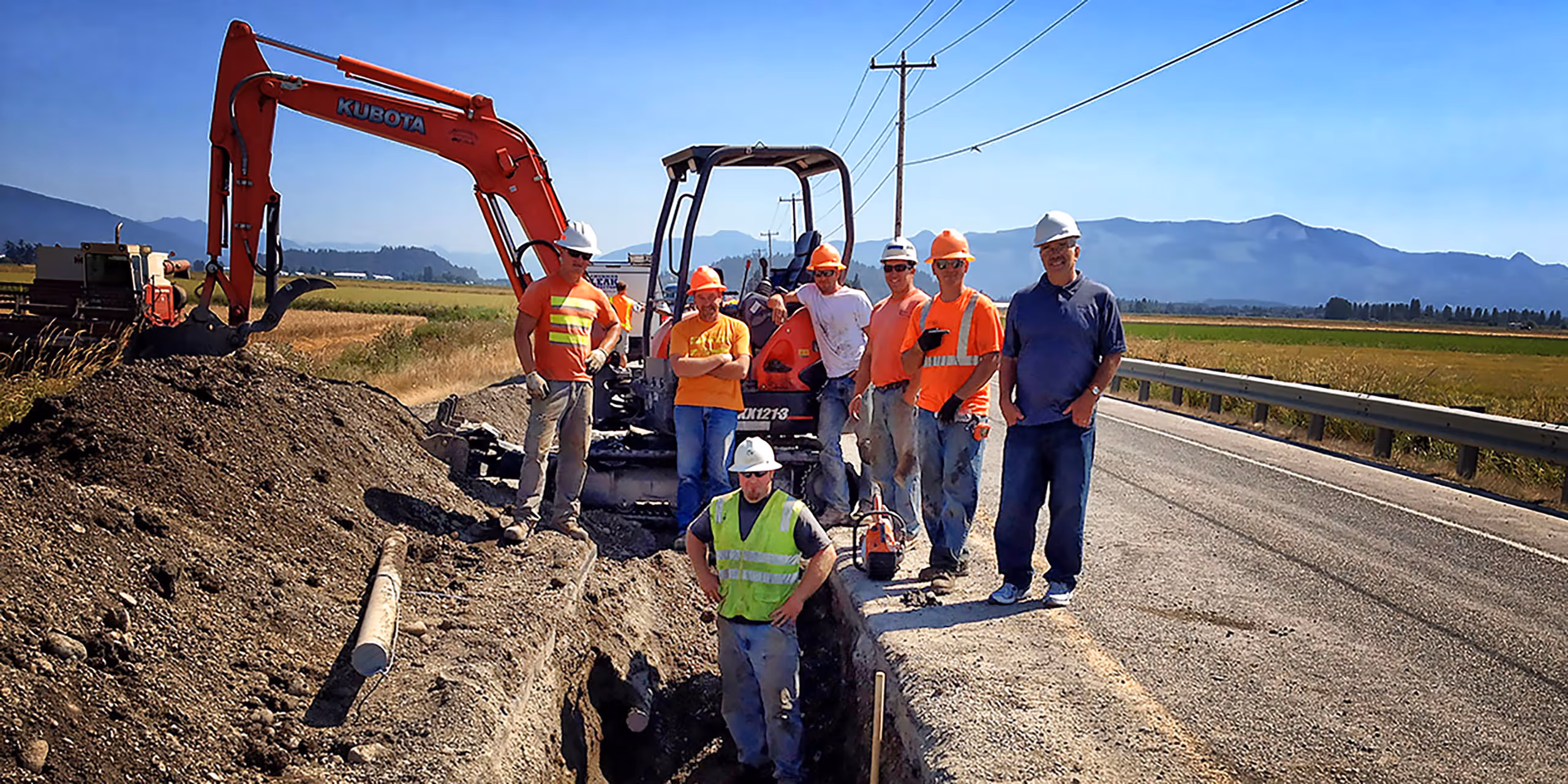 A group of seven construction workers stands near a trench on the side of a rural road. They are wearing safety vests and helmets, with construction equipment, including an excavator, visible in the background. The landscape features fields and utility poles.