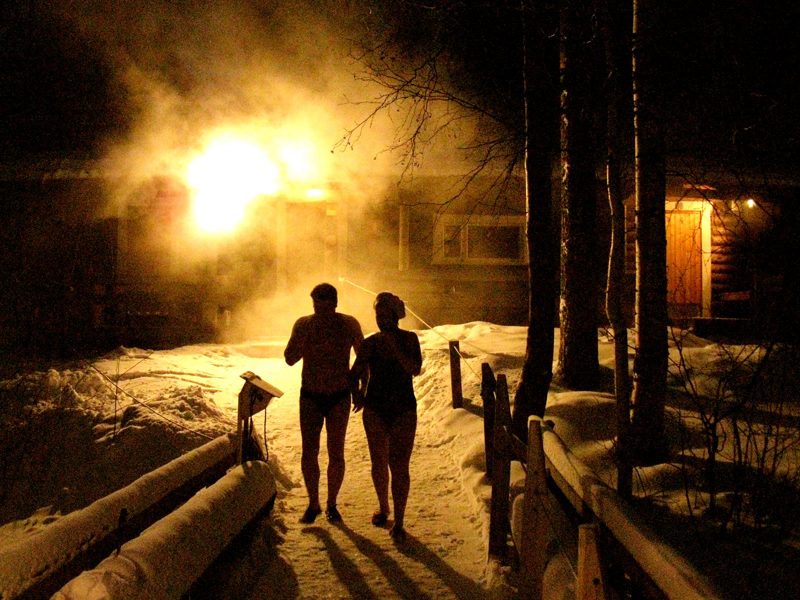 A couple walking from a sauna to a hole cut in the ice