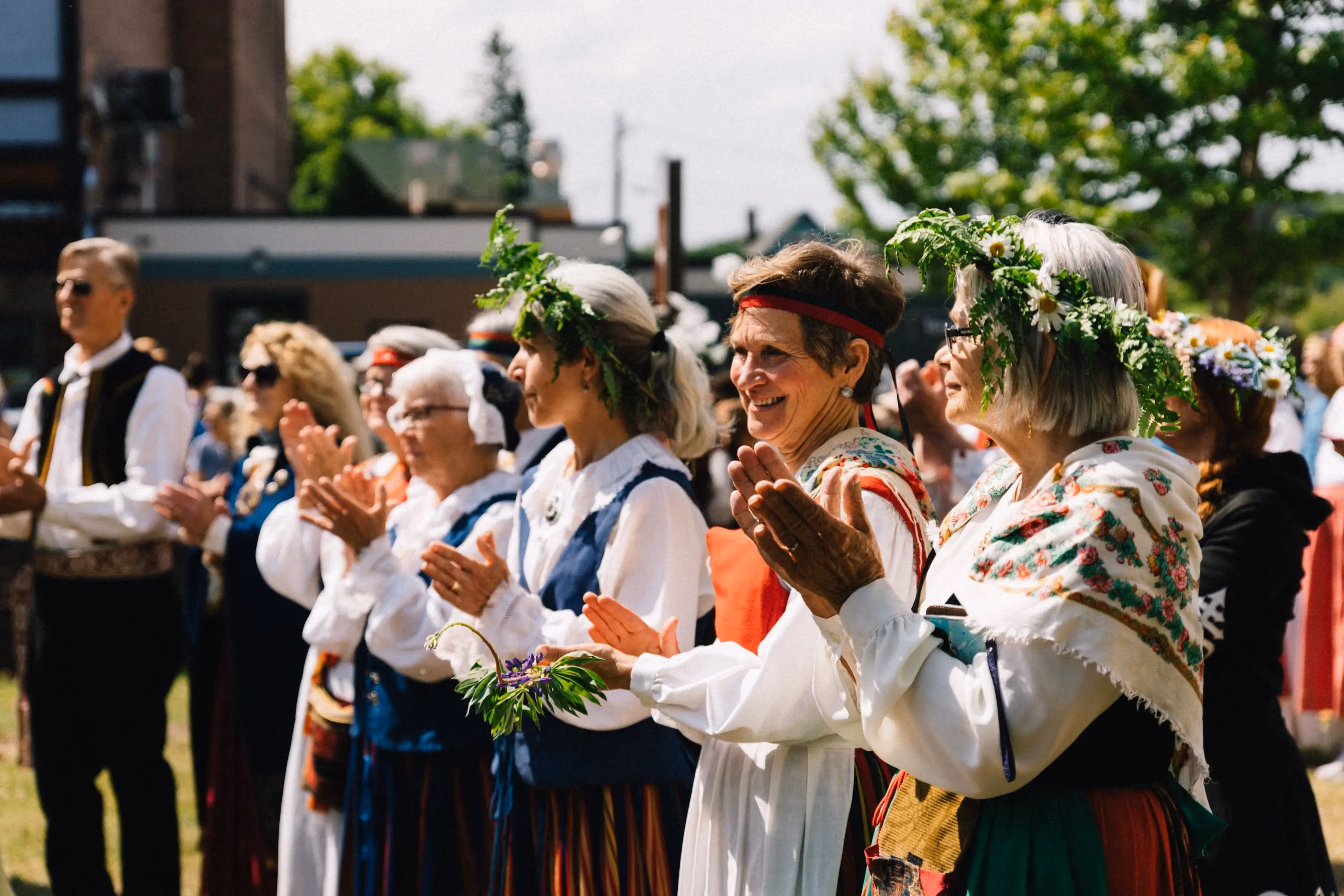 A row of people in Nordic folk dress applaud outdoors in sunshine, several wearing midsummer wreaths of greenery and daisies.