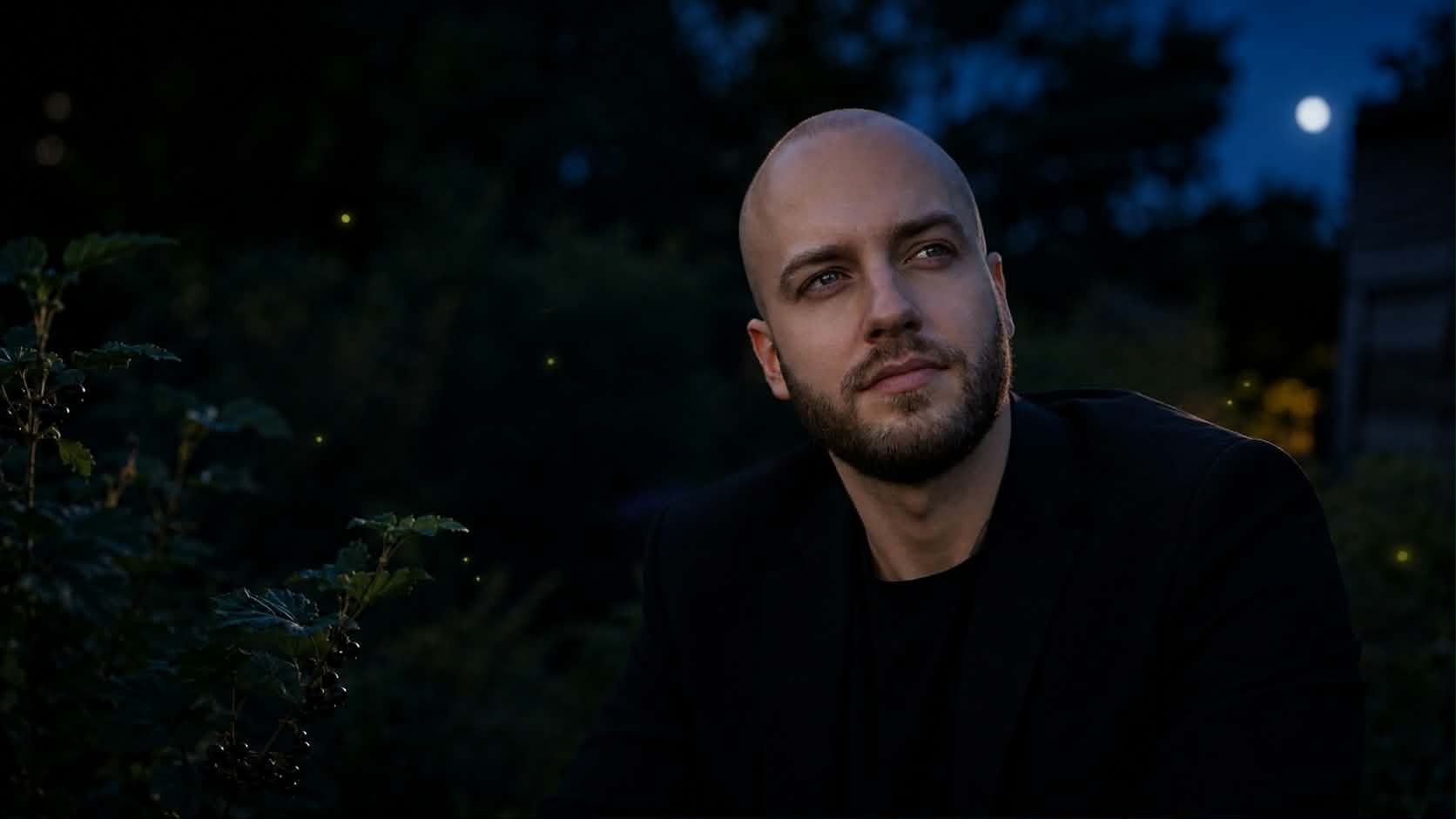 A man in a dark jacket sits outdoors at dusk, a full moon rising above the trees behind him.