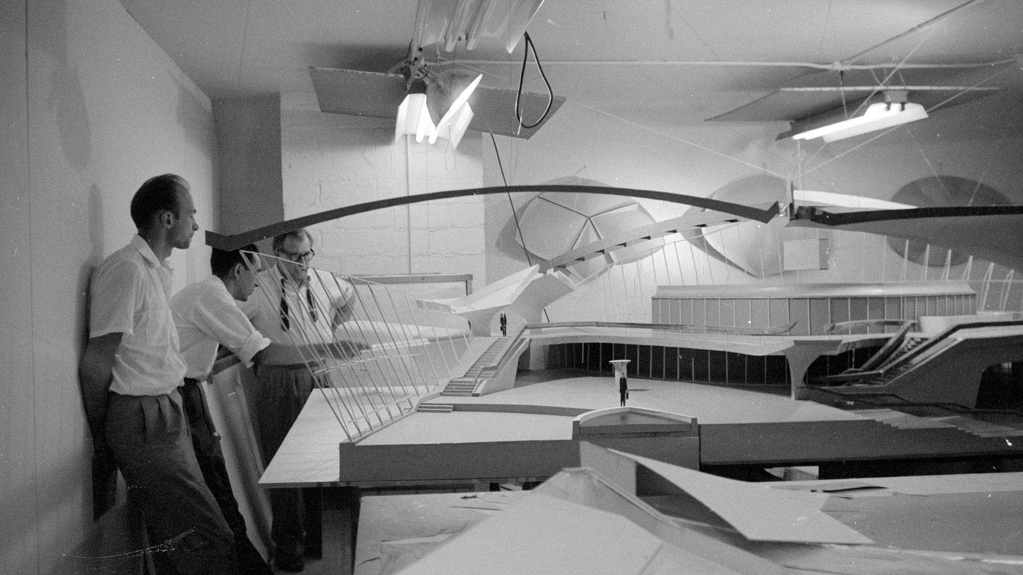 Three men in a mid-century studio study a large architectural model with sweeping curved roof forms.