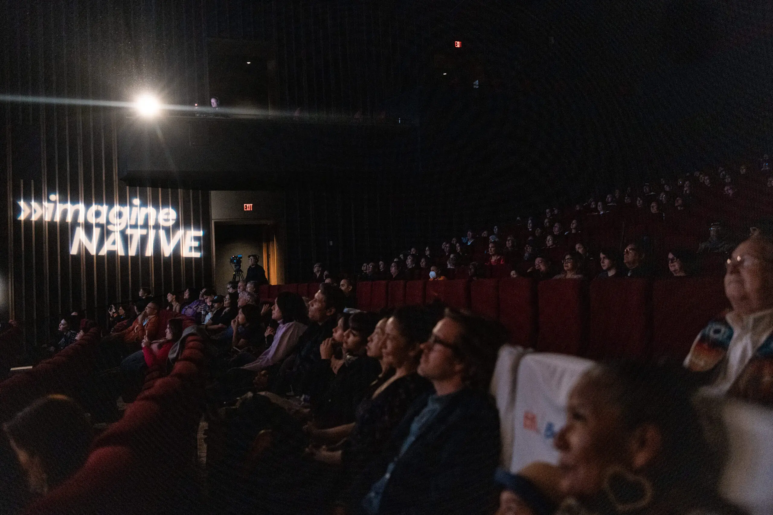 A seated audience in a darkened cinema, with the imagineNATIVE logo lit on the wall beside the screen.
