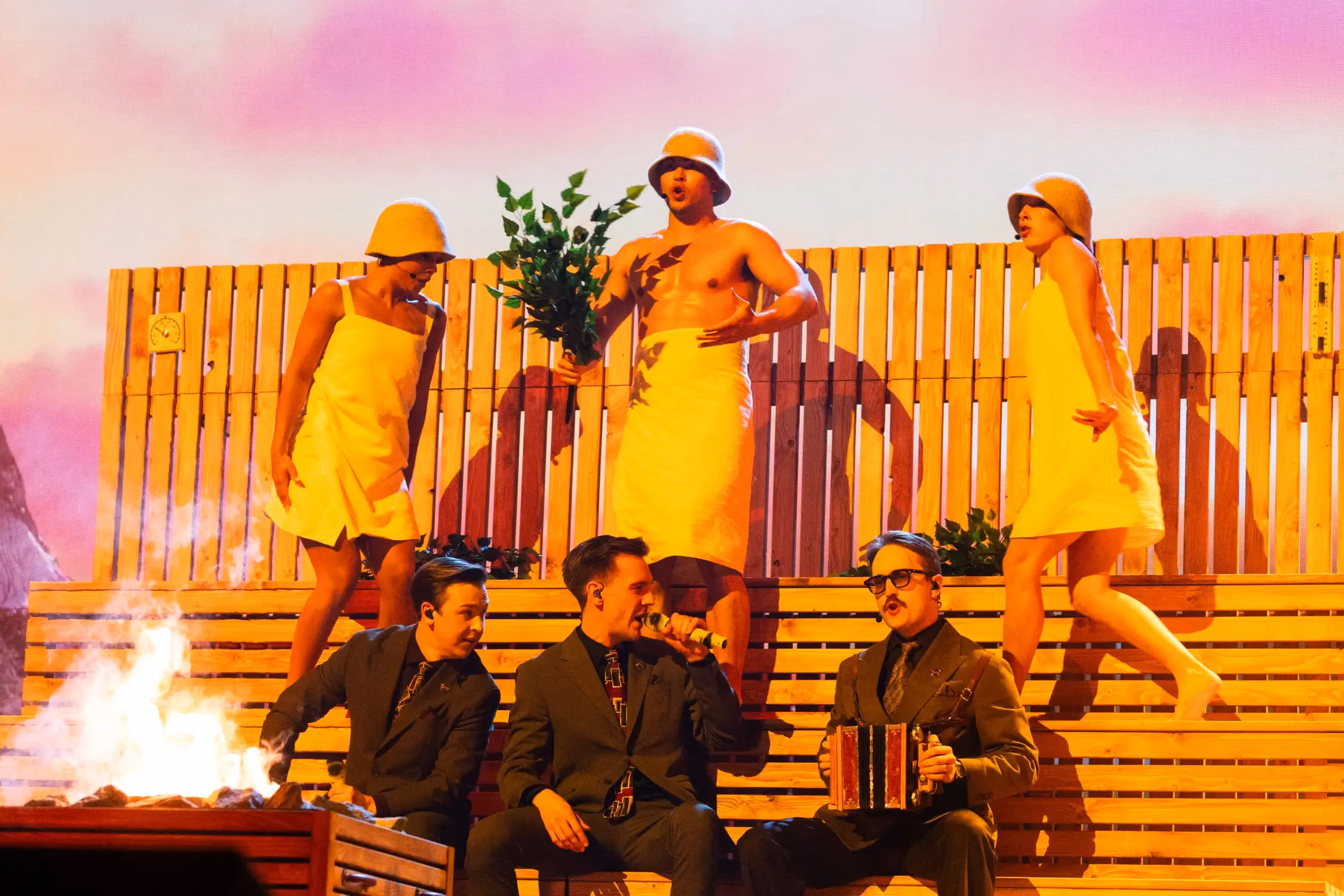 Performers in towels and felt hats on a sauna-themed stage set, with three singers in suits seated below and a stove fire burning.