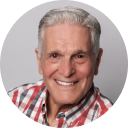 Smiling older man with gray hair wearing a red and blue plaid shirt and white t-shirt.