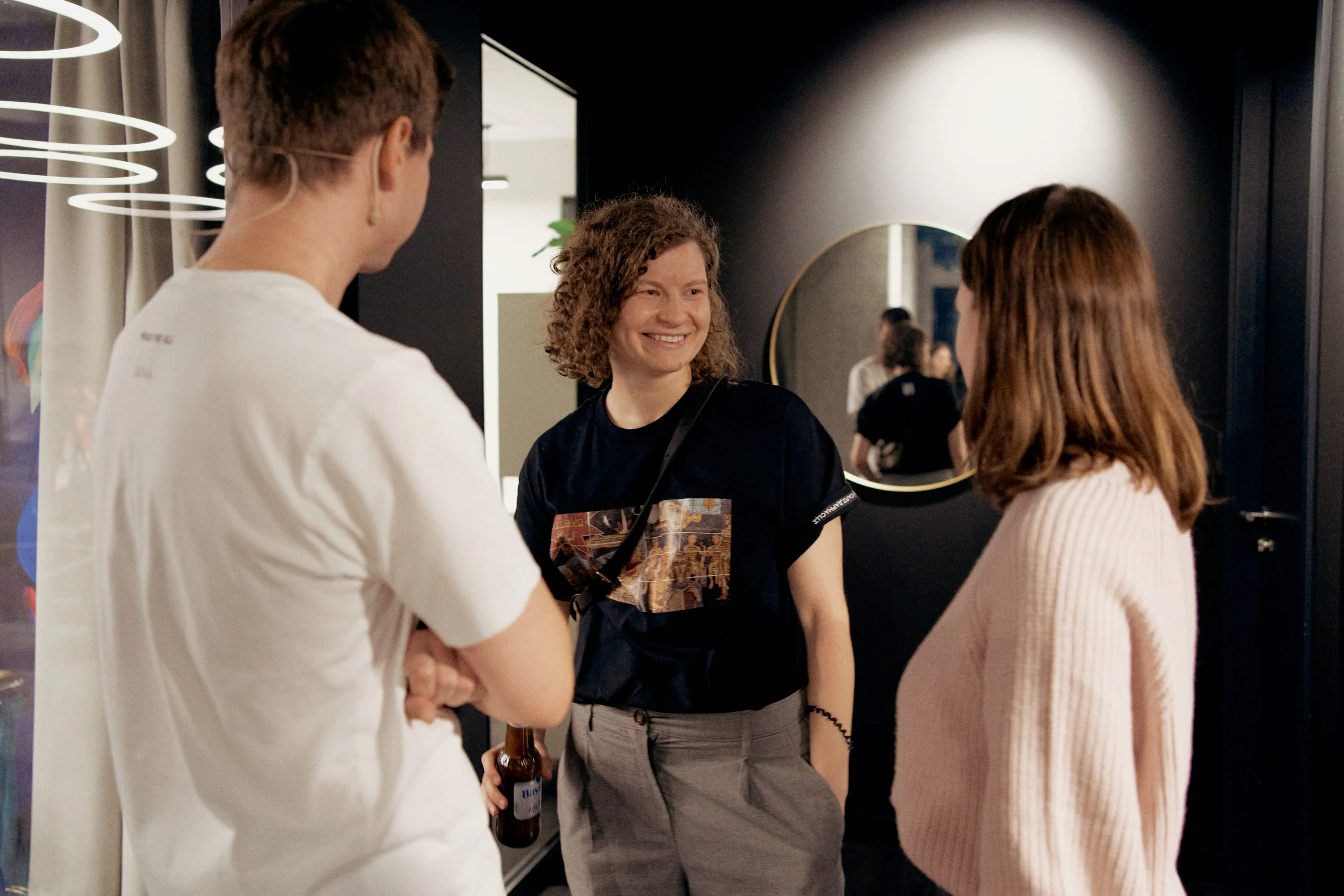 Three people standing indoors engaged in conversation; one person with curly hair in a black shirt and gray pants is smiling and holding a bottle, another in a light pink sweater, and the third person with short brown hair in a white t-shirt facing them.