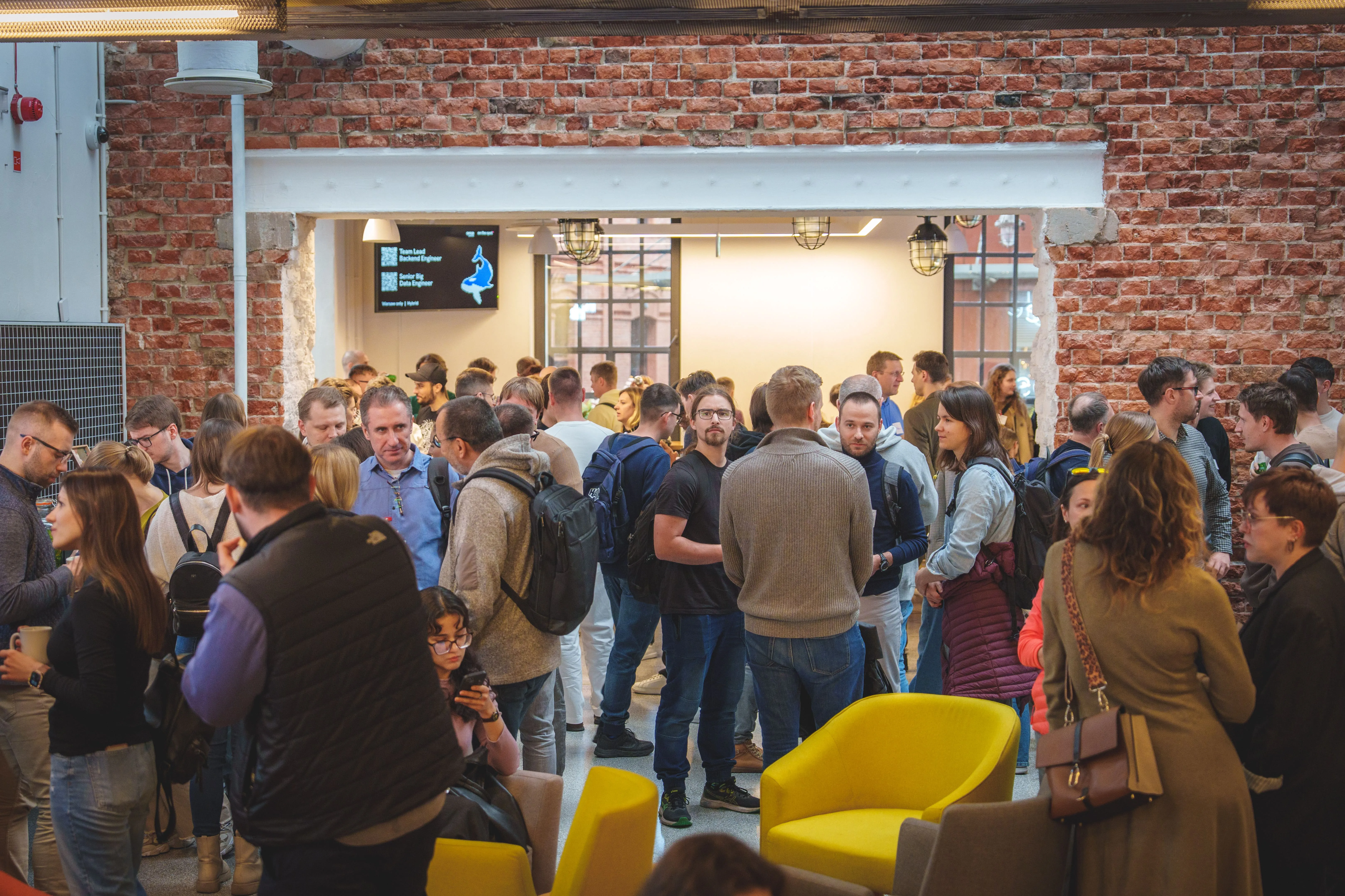 Large group of people socializing inside a modern room with exposed brick walls and yellow chairs, some wearing backpacks and engaged in conversation.