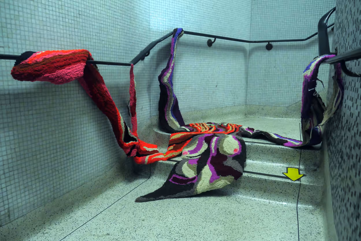 Photo of an installation with brightly coloured woven forms draped over handrails in a tiled hallway.