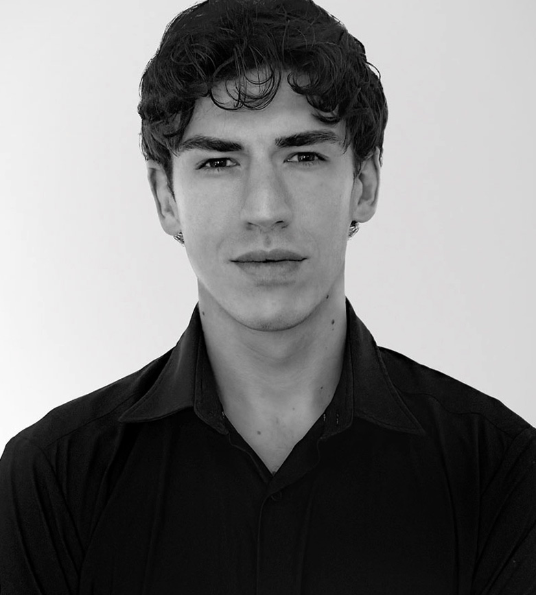Black and white portrait of a young man with dark curly hair wearing a black collared shirt, looking directly at the camera against a plain background.
