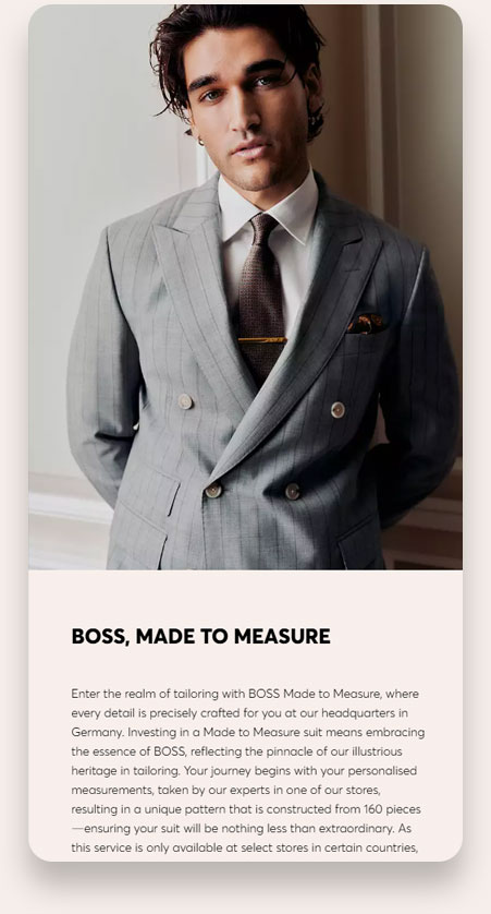 A man with dark hair wearing a gray, double-breasted pinstripe suit, white shirt, dark tie with a tie bar, and a patterned pocket square, posing indoors against a light wall.
