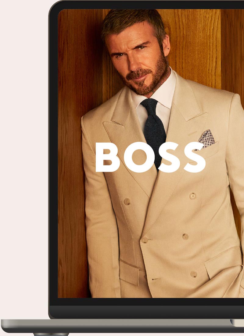 A man with short dark hair and a beard wearing a beige double-breasted suit, white shirt, and dark tie posed in front of a wooden background, with the word BOSS in bold white letters across the image.