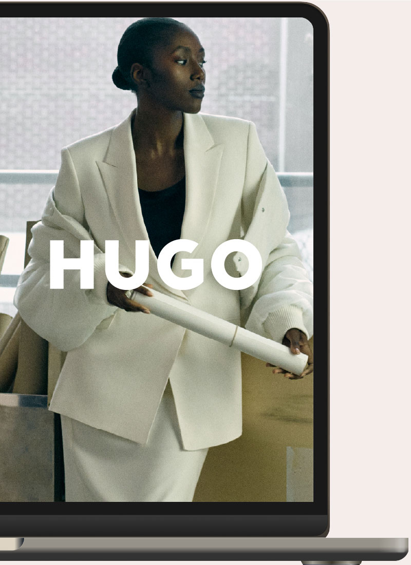A woman wearing a white blazer and skirt with a black top, holding a rolled-up paper, standing indoors near a window, with the word 'HUGO' overlaid in white text.