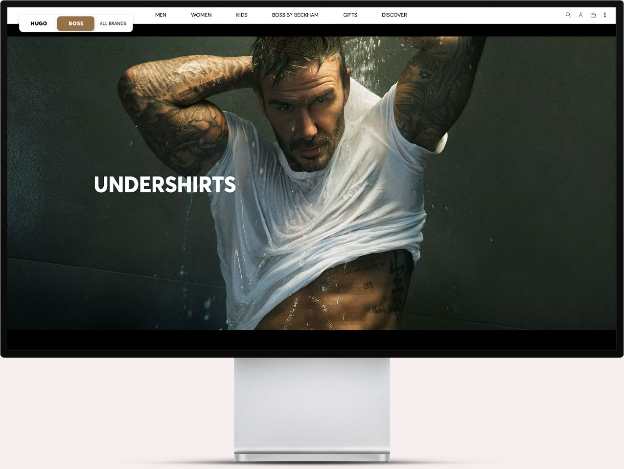 A tattooed man wearing a wet white undershirt, lifting it up with water pouring over him, displayed on a computer monitor with a website menu showing categories like Men, Women, Kids, Boss by Beckham, Gifts, and Discover.