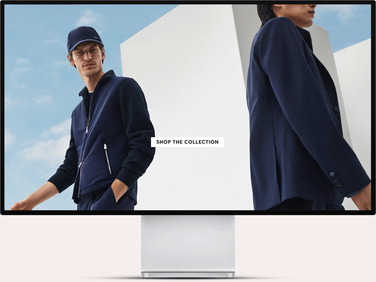 Computer monitor displaying a fashion ad with two male models wearing navy blue outerwear against a sky and modern white background, with text 'SHOP THE COLLECTION' in the center.