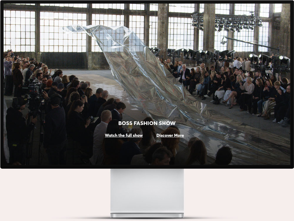 A fashion show runway with a large, reflective silver fabric installation that rises dramatically from the floor; the runway is surrounded by seated audience on both sides and large windows in the background.
