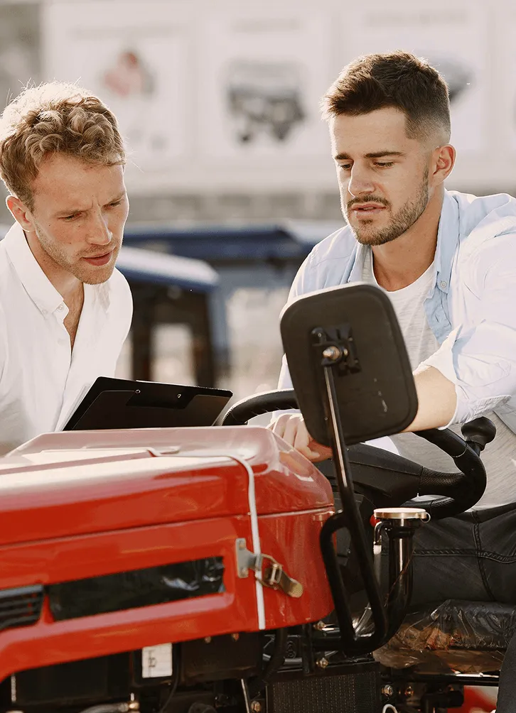 Two men inspecting and discussing a red tractor, one holding a clipboard and the other pointing at controls.
