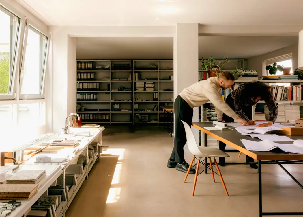 Two professionals reviewing large blueprints on a table in a bright, modern office with shelves of books and materials.