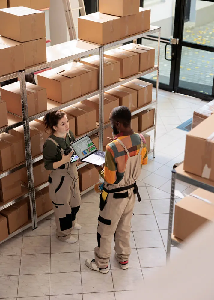Two warehouse workers in overalls discussing while checking inventory on a laptop surrounded by cardboard boxes on metal shelves.