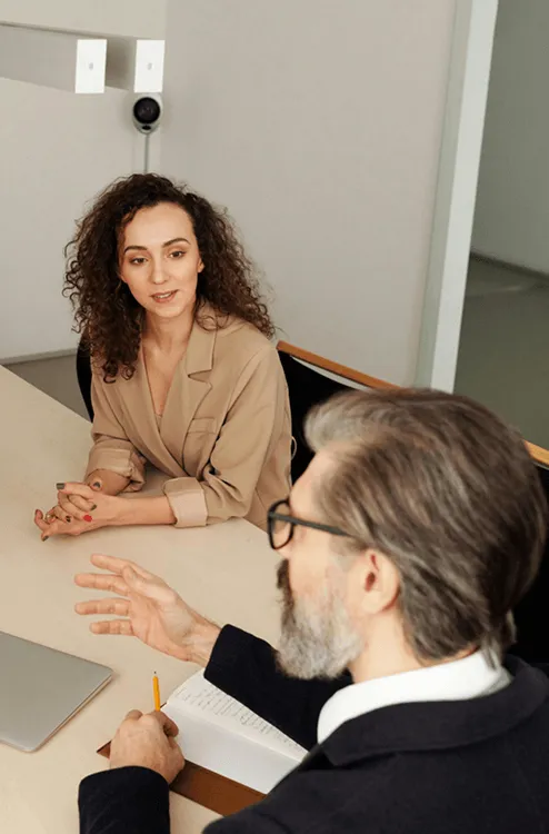 Professional man with glasses talking to a woman in a tan blazer during a meeting at a table with a laptop and notebook.