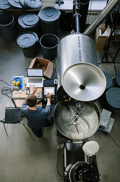 Person operating a large industrial coffee roaster with a laptop and control devices on a nearby table in a roasting facility.