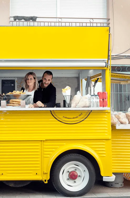 Man and woman smiling and serving food from a bright yellow food truck window with snacks and drink bottles displayed.