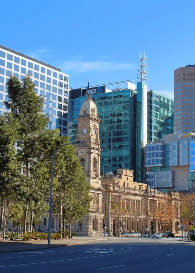 Historic sandstone clock tower building with modern glass skyscrapers behind and trees in the foreground under a clear blue sky in Adelaide.