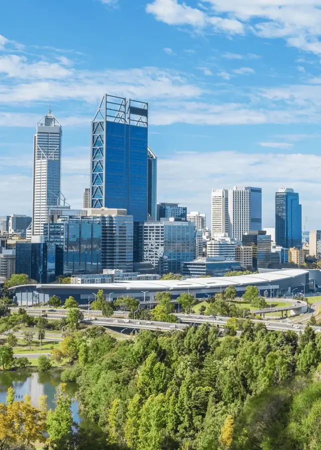 Skyline of Perth city with modern skyscrapers under a partly cloudy blue sky, green trees and a water body in the foreground.