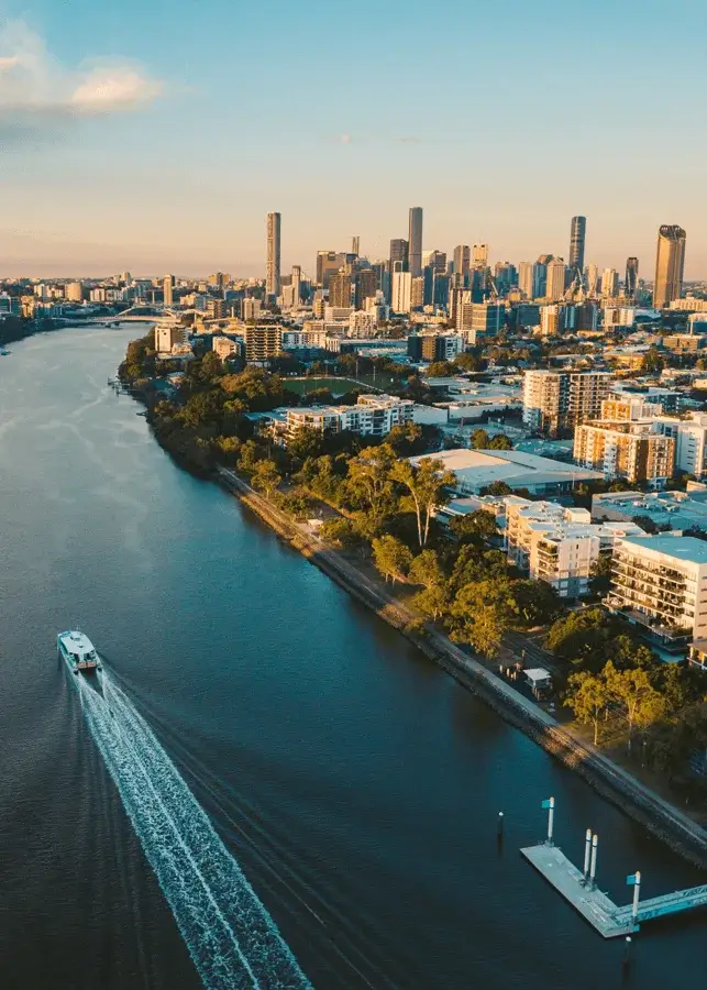 Aerial view of a river with a boat leaving a wake, bordered by lush green trees and urban buildings, with a city skyline in the background under a clear sky at sunset.