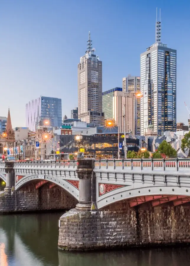 View of a bridge over the Yarra River with Melbourne skyline and skyscrapers in the background during early evening.