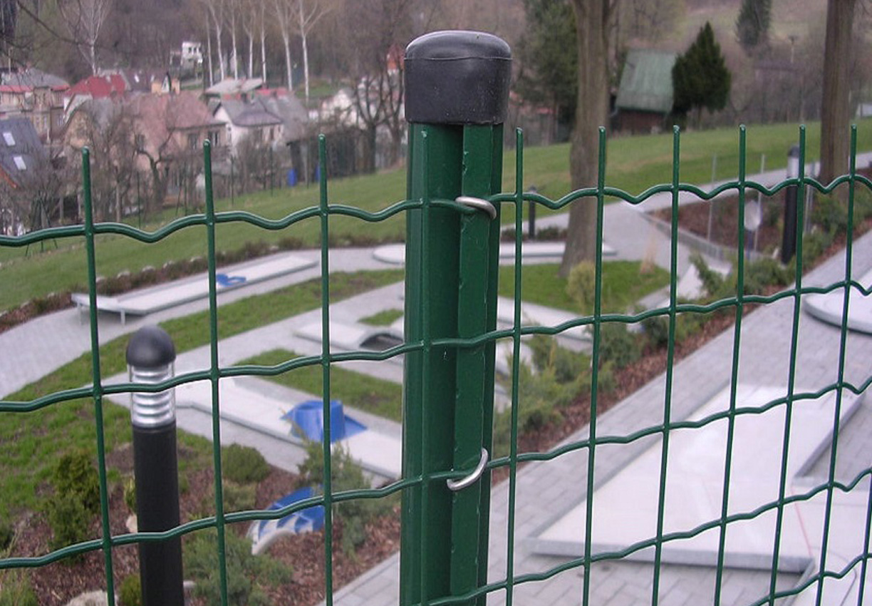 Zöld fémrácsos kerítés előtérben, mögötte park, lámpa és házak láthatók.