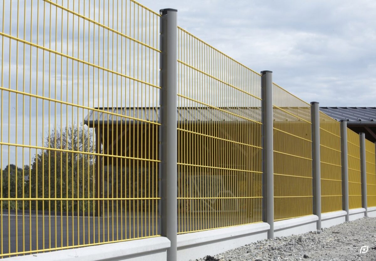 Sárga fémrács kerítés szürke oszlopokkal és beton alapzattal, épület mögött.