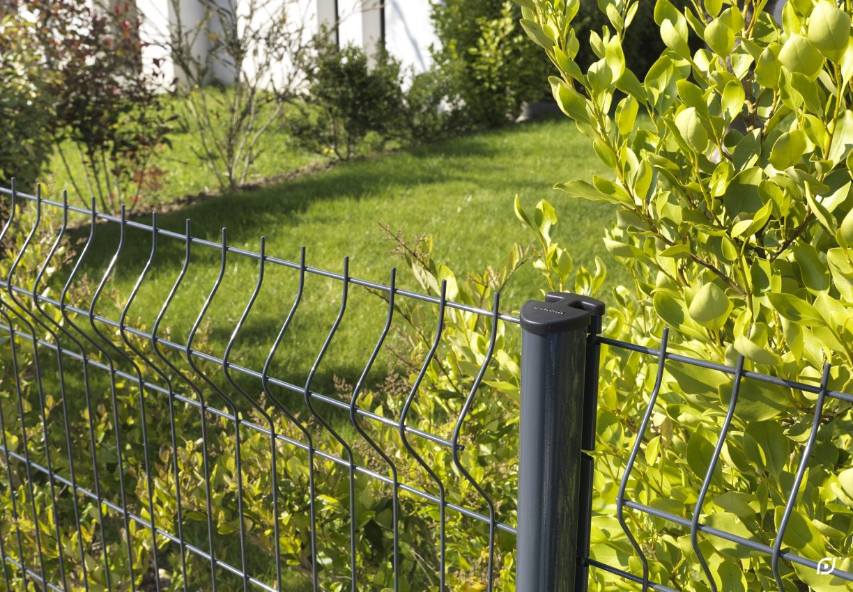 Fémkerítés sötétszürke oszloppal és zöld levelekkel egy napsütötte kertben.