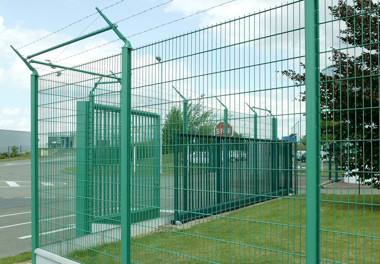 Zöld, fém biztonsági kerítés szögesdróttal és kapuval ipari területen.
