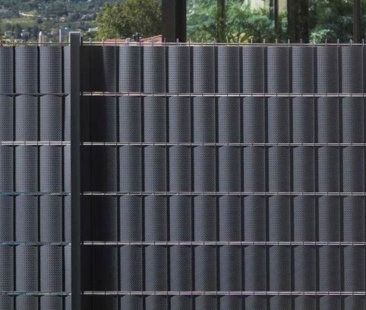 Sötétszürke fém kerítés vízszintes és függőleges rudakkal, háttérben fák és épületek.