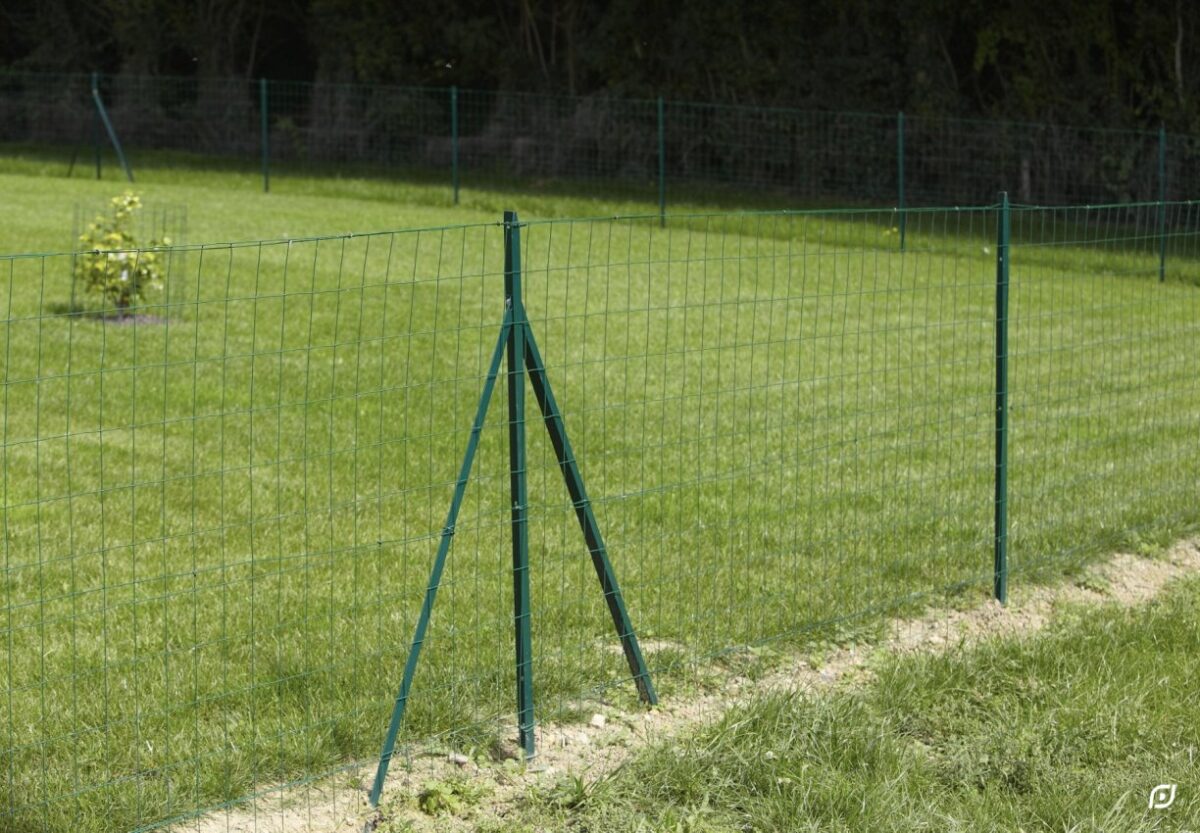 Zöld fémkerítés támasztó oszlopokkal egy füves kert mellett.