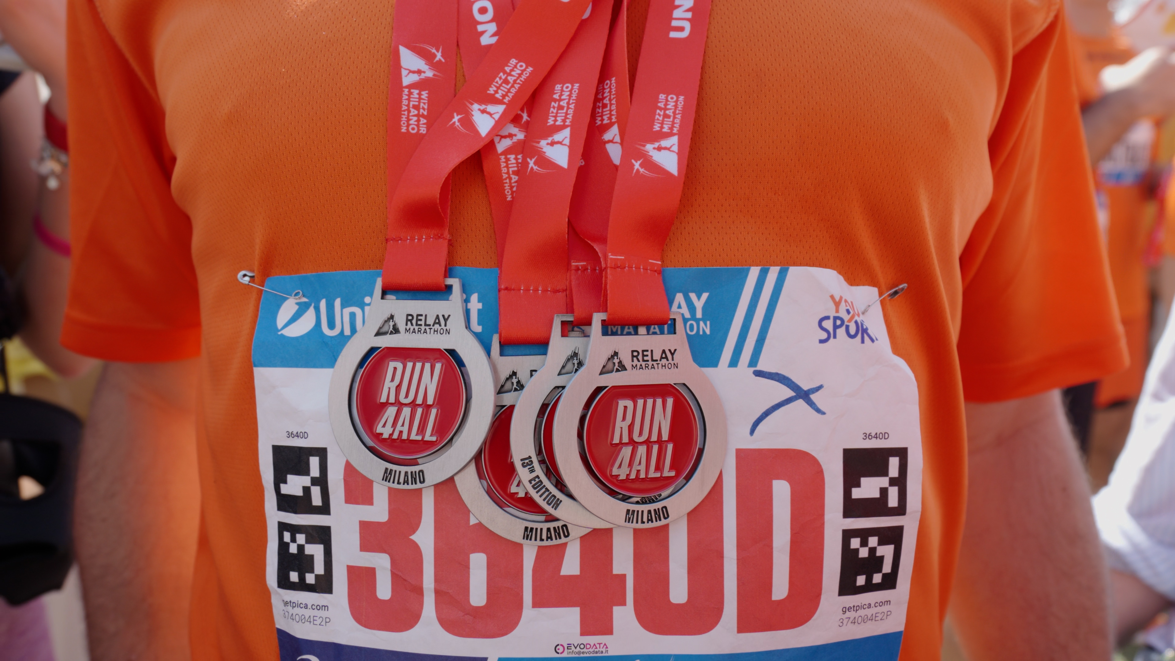 Gruppo di giovani sorridenti con medaglie, alcuni indossano magliette arancioni con la scritta YouSport, festeggiano all'aperto dopo una gara.