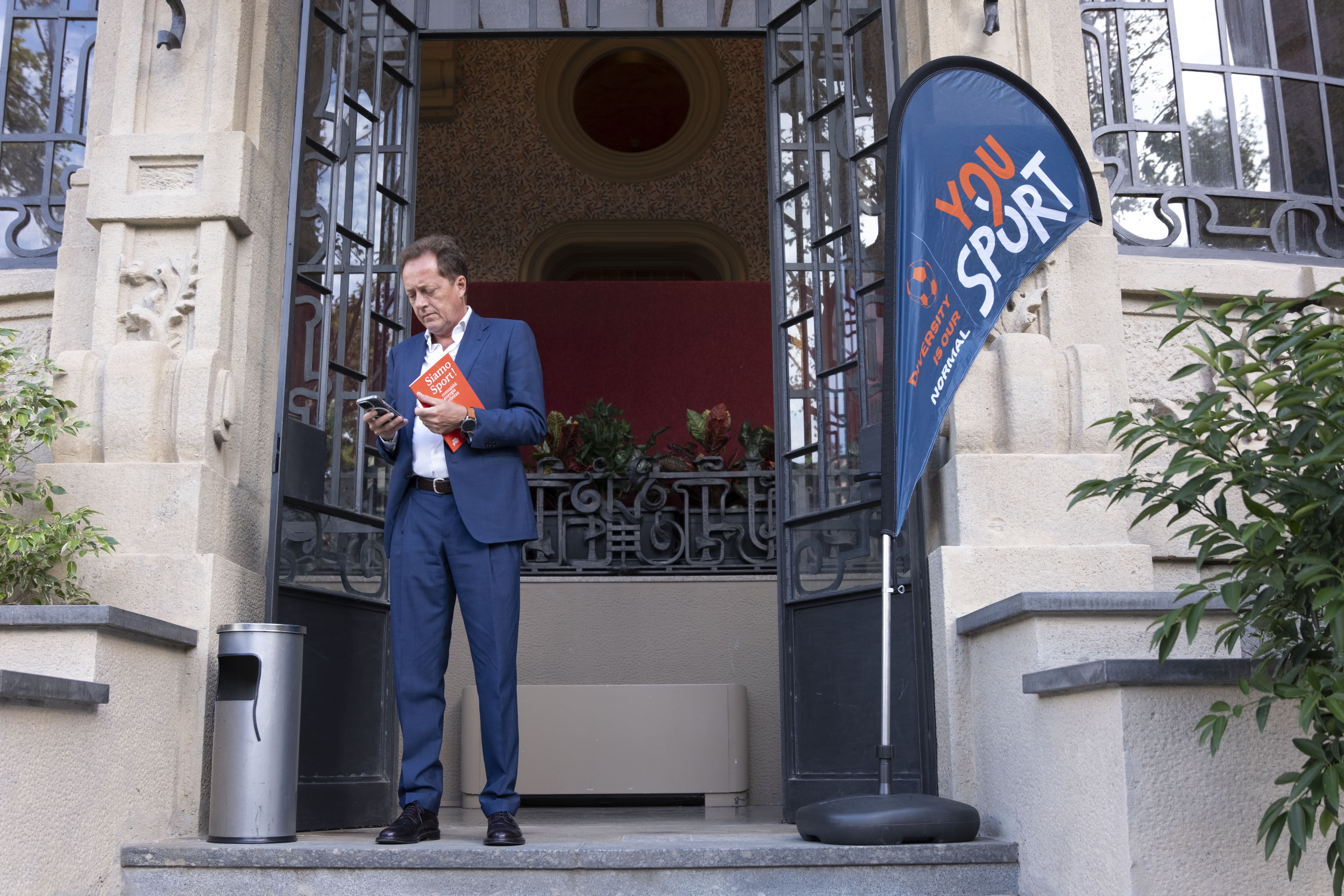 Gruppo di persone che partecipano a una corsa indossando magliette arancioni con scritte You Sport, alcune fanno il segno della vittoria e tengono uno striscione con la scritta 'Diversity is our normal'.