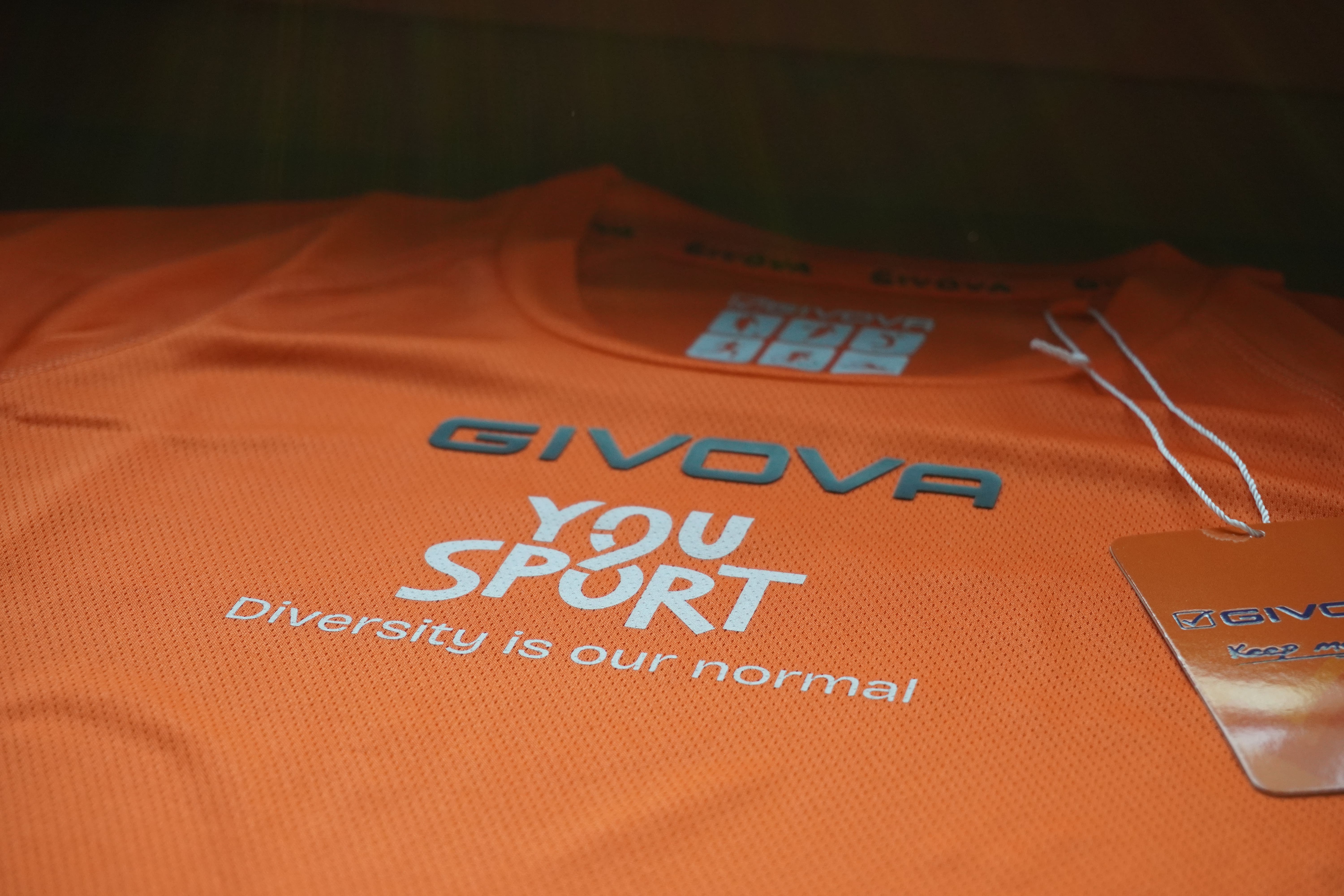 Due donne sorridenti con magliette arancioni etichettate 'You Sport' durante una maratona di staffetta, con volti truccati, circondate da altri corridori.