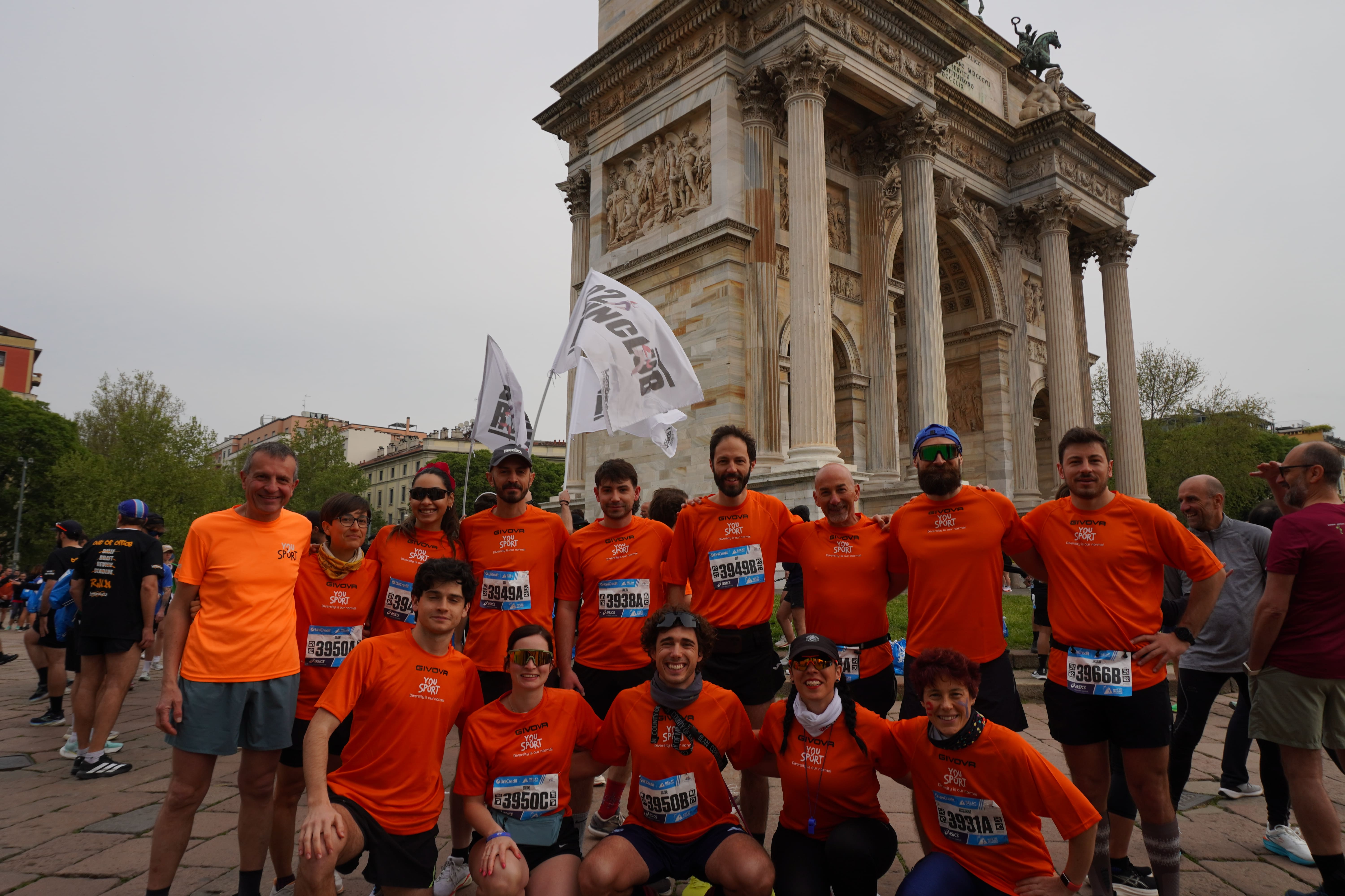 Due donne sorridenti con magliette arancioni etichettate 'You Sport' durante una maratona di staffetta, con volti truccati, circondate da altri corridori.