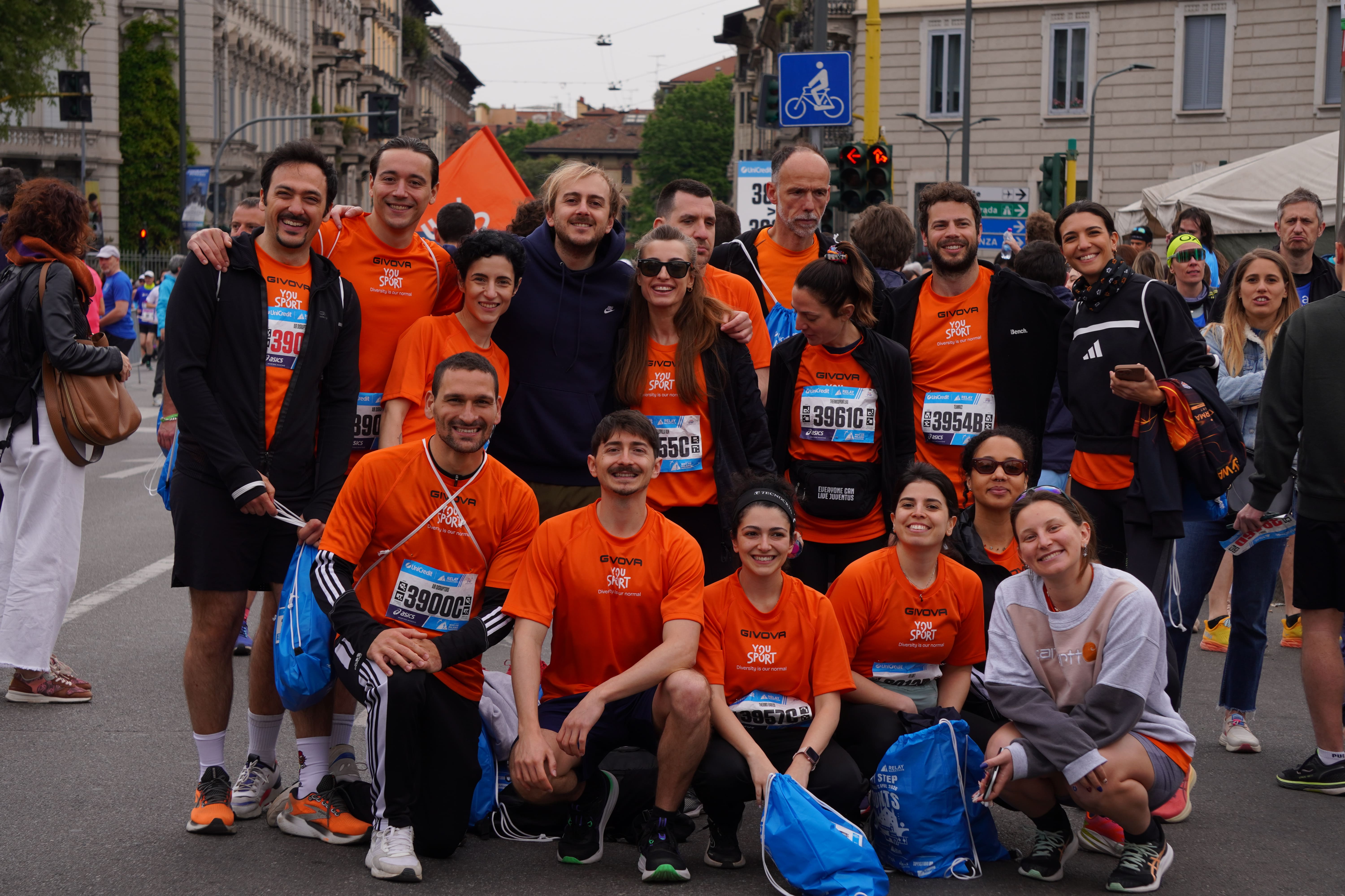 Gruppo di persone sorridenti con magliette arancioni YouSport, partecipanti a un evento all'aperto in una piazza storica con edifici e colonne.